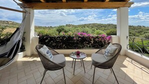 Eine überdachte Terrasse mit zwei Korbstühlen und einem kleinen Tisch, mit Blick auf eine üppige Landschaft mit Hügeln und blauem Himmel.