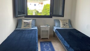 Ein kleines Zimmer mit zwei Betten, einem Fenster mit geöffneten Rollläden, einem Nachttisch und Blick auf ein Gebäude und Grünflächen.
