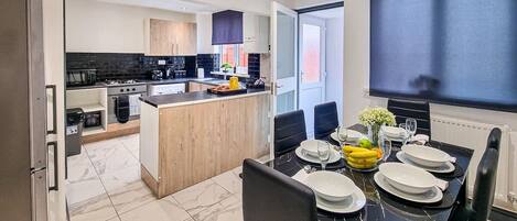 A modern kitchen with a central island, a dining area with a black marble table, and a large TV on the wall.