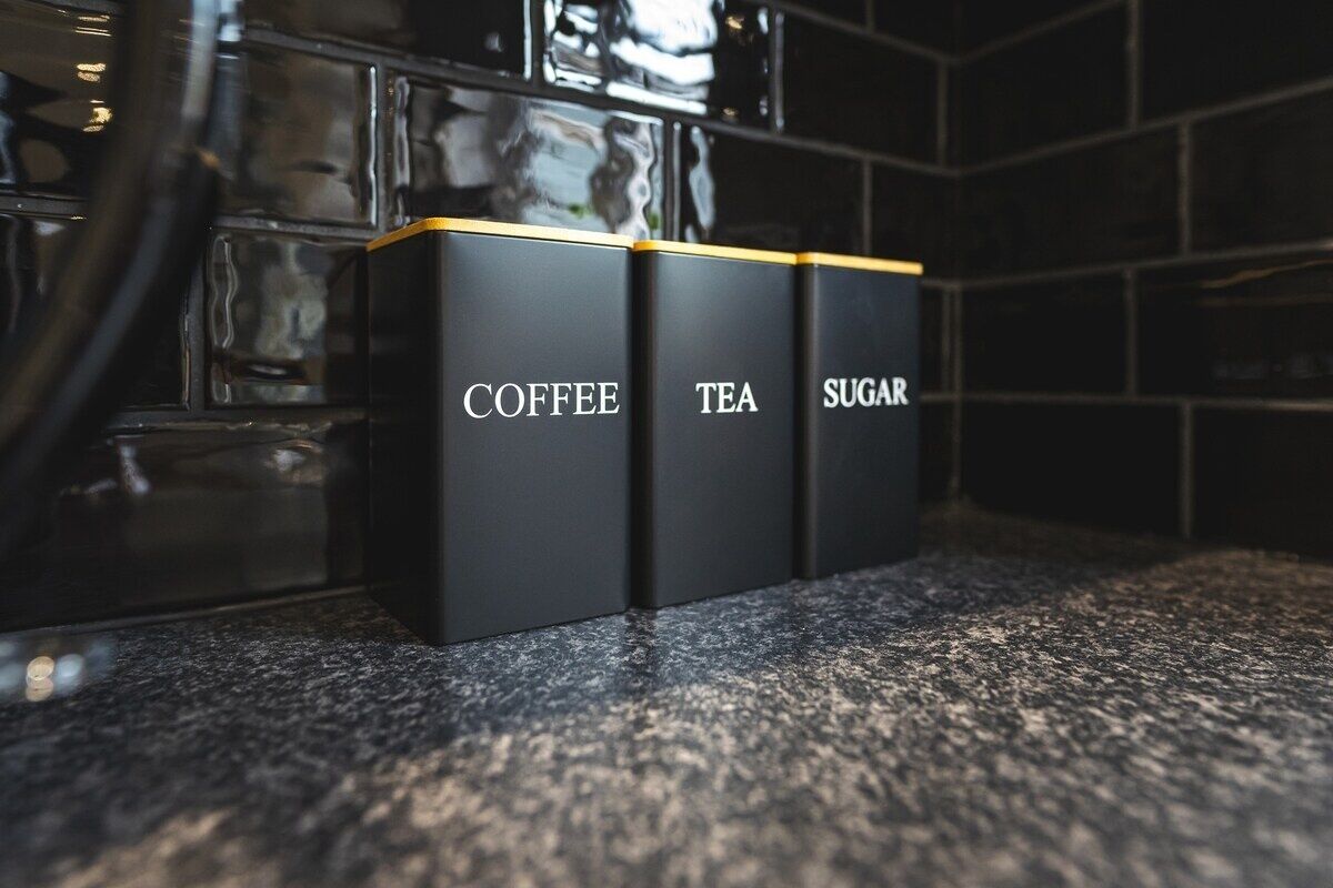 Drei schwarze Behälter mit den Aufschriften Kaffee, Tee und Zucker auf einer Arbeitsplatte.