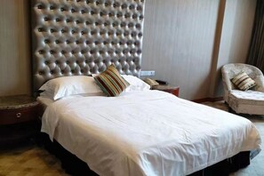 Ett hotellrum med en stoppad sänggavel, en säng med vita sängkläder, en randig kudde, ett sängbord med en telefon och en stol med en randig sittkudde.