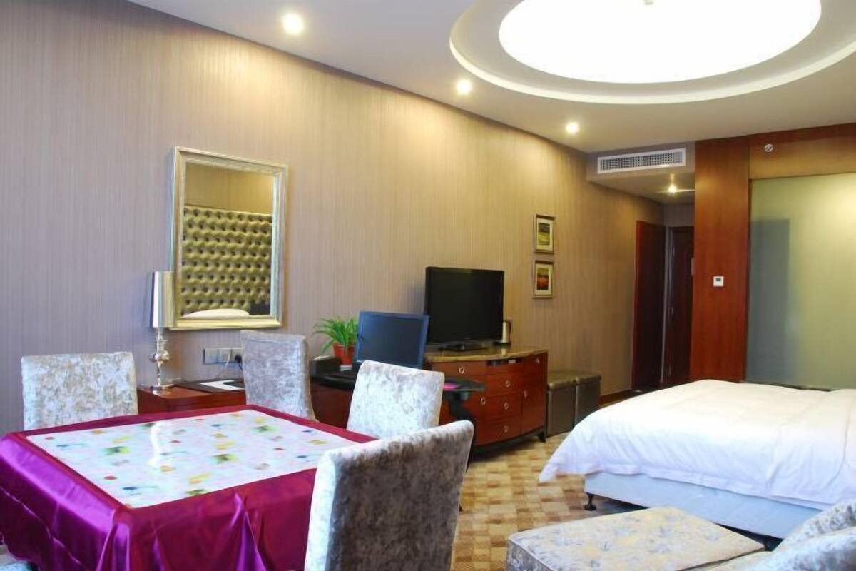 Ein Hotelzimmer mit einem Bett, einem Fernseher, einem Schreibtisch mit Stuhl, einem Esstisch mit rotem Tischläufer und einem großen Spiegel.