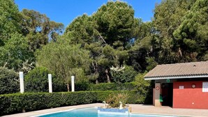 A clear blue pool surrounded by lush greenery and a red building with a tiled roof. - Appartement avec jardin - Cavalière (Le Lavandou)