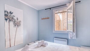 A bedroom with a bed, a window with a view, a radiator, and a wall-mounted artwork. - Appartement avec jardin - Cavalière (Le Lavandou)