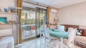 A room with a dining table, chairs, a sofa, a radiator, a lamp, and a window with curtains. - Appartement avec jardin - Cavalière (Le Lavandou)