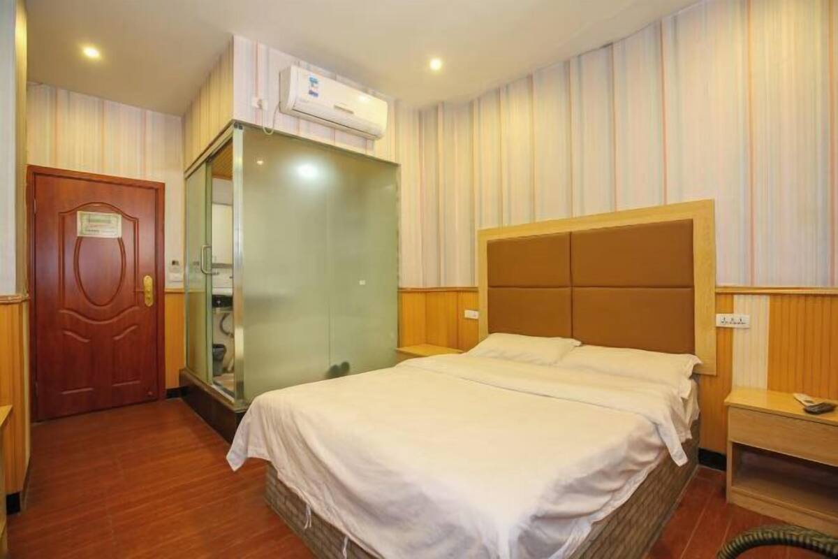 Habitación de hotel con cama, puerta de madera, baño con cabina de vidrio y aire acondicionado instalado en la pared.