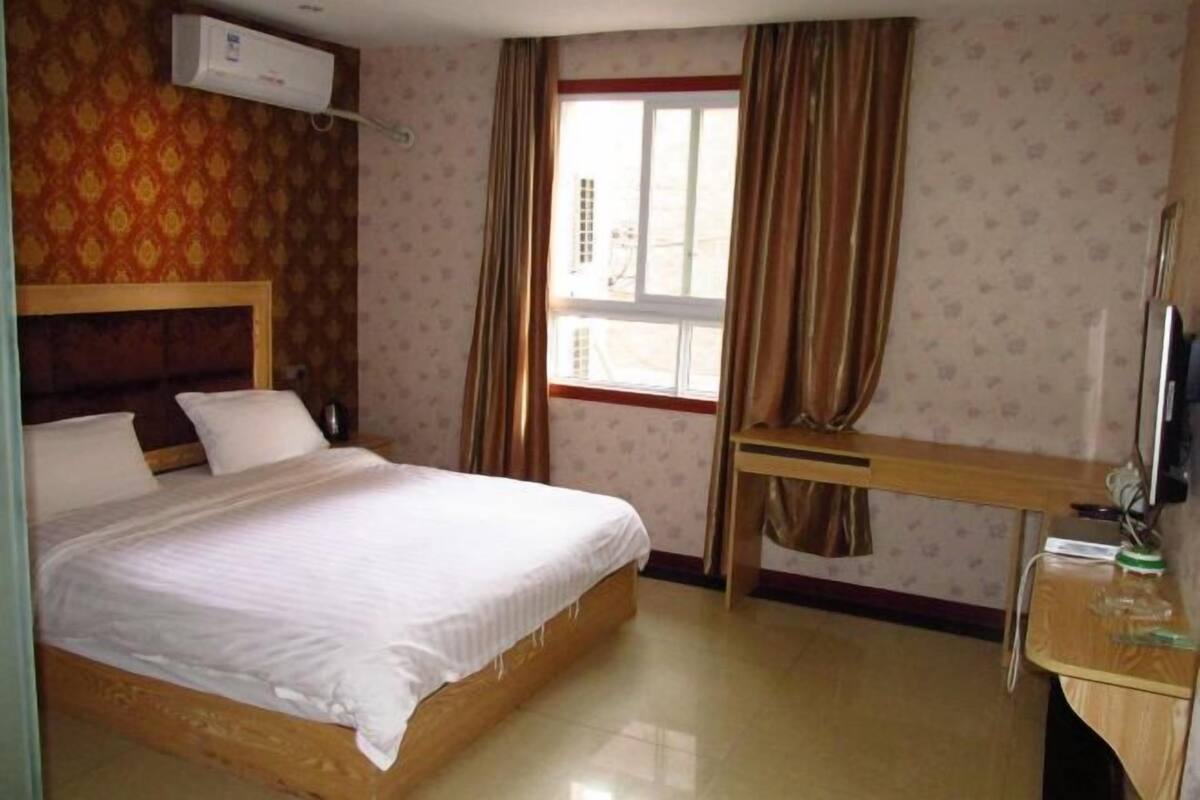 Una habitación de hotel con cama, escritorio, silla, unidad de aire acondicionado y ventana con cortinas.
