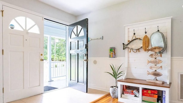 Una entrada con suelo de madera, una puerta blanca, una planta en maceta y una estantería con libros y peces decorativos.