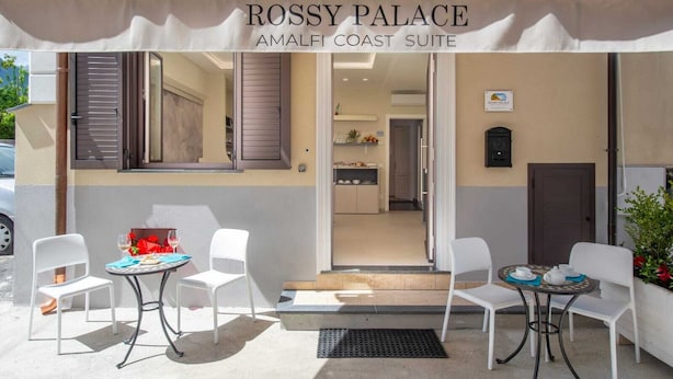 Esterno del Palazzo Rossy, con una zona salotto all'aperto coperta, due tavoli e sedie, vista sull'interno e un cartello che indica la location come Amalfi Coast Suite.