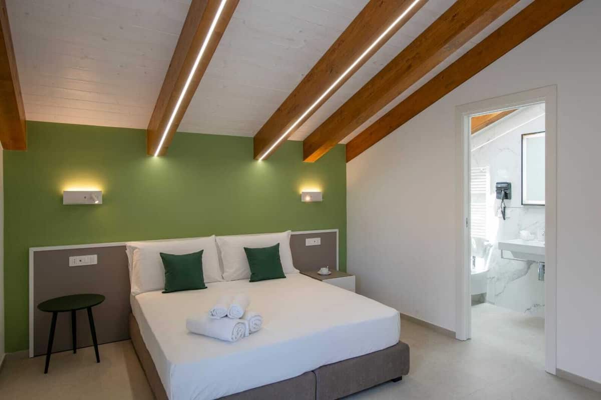 Une chambre d’hôtel moderne avec un grand lit, des murs verts, des poutres en bois et une salle de bain visible à travers une porte ouverte.