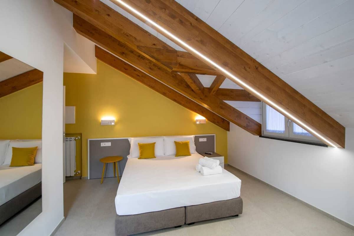 Une chambre avec un plafond en pente, des poutres en bois, un lit avec une literie blanche, deux tables de chevet et un petit tabouret.
