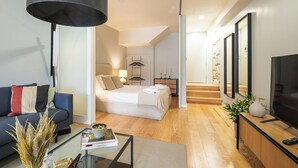 A modern living room with a sofa, coffee table, and a TV. A bedroom with a bed and nightstand is visible through an open door.