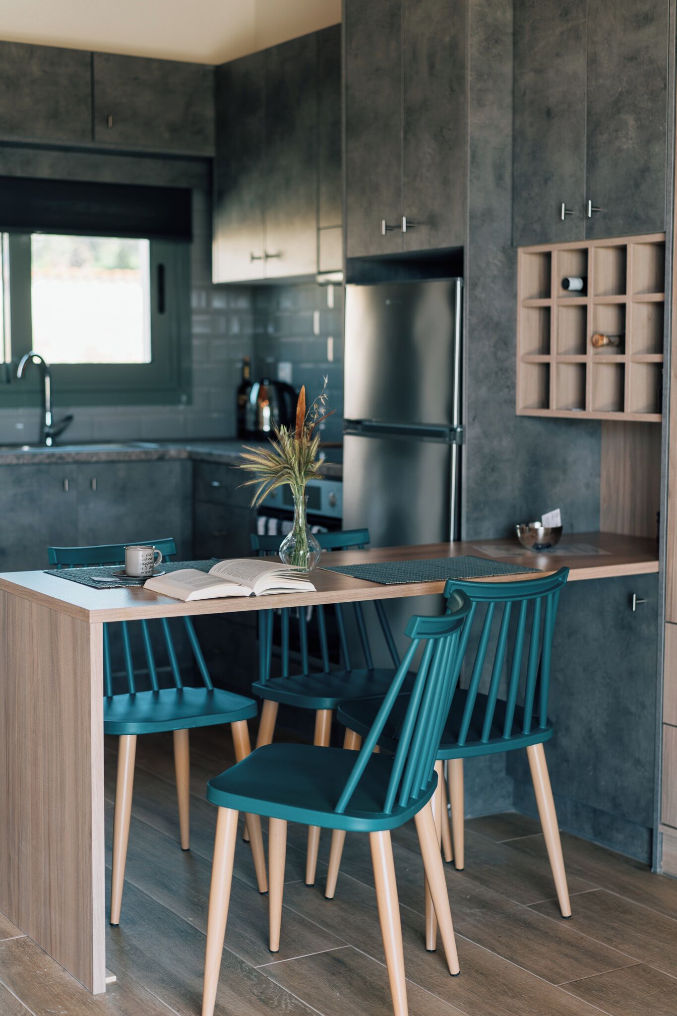 Una cocina moderna con armarios oscuros, una isla de madera, sillas color turquesa y una pequeña mesa de comedor.