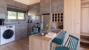 A modern kitchen with grey cabinets, a built-in washing machine, and a dining area with a wooden table and teal chairs. - Apolithon Premium Houses (Lesvos)