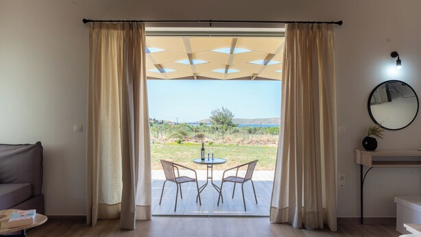 A room with a sliding glass door leading to an outdoor area, a round table with two chairs, a sofa, a small table with a bottle, a mirror, and a wooden desk. - Apolithon Premium Houses (Lesvos)