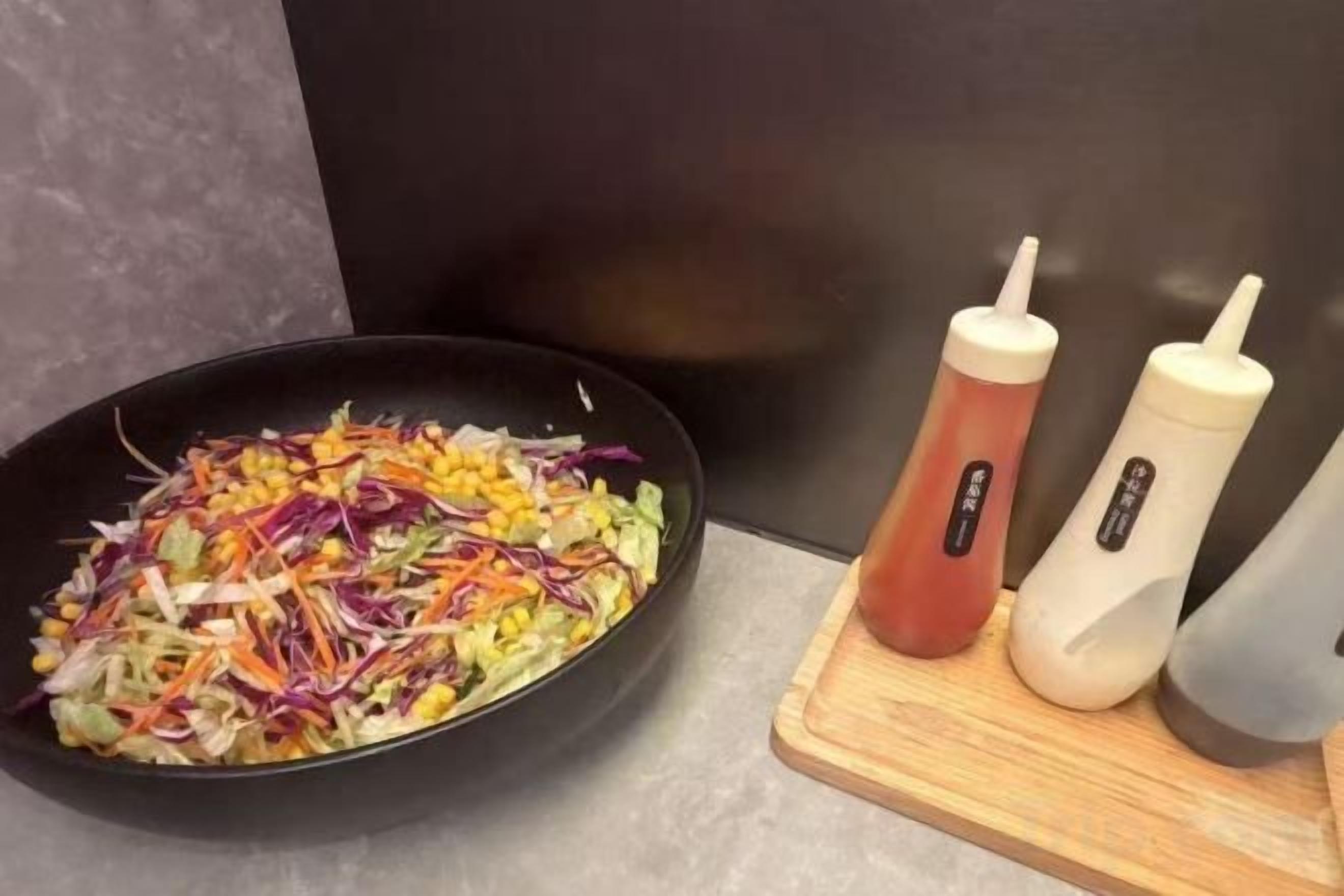 A bowl of salad with various vegetables and two condiment bottles on a wooden board.