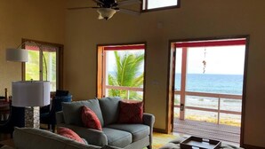 A living room with a gray sofa, red cushions, a lamp, and a view of the beach through large windows. - The Whole Hibiscus - Pool & Ocean Front! (Utila)
