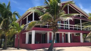 A two-story building with a red exterior and white columns, surrounded by palm trees on a sandy ground. - The Whole Hibiscus - Pool & Ocean Front! (Utila)