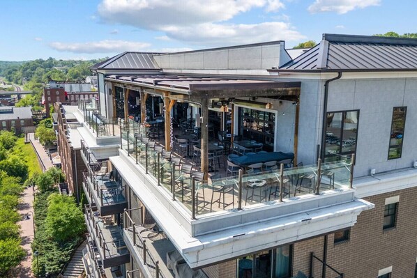 A rooftop bar with outdoor seating and a glass railing, overlooking a cityscape.