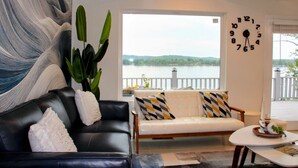 A living room with a black leather sofa, a white sofa with patterned cushions, a large abstract painting, a green plant, and a clock on the wall.