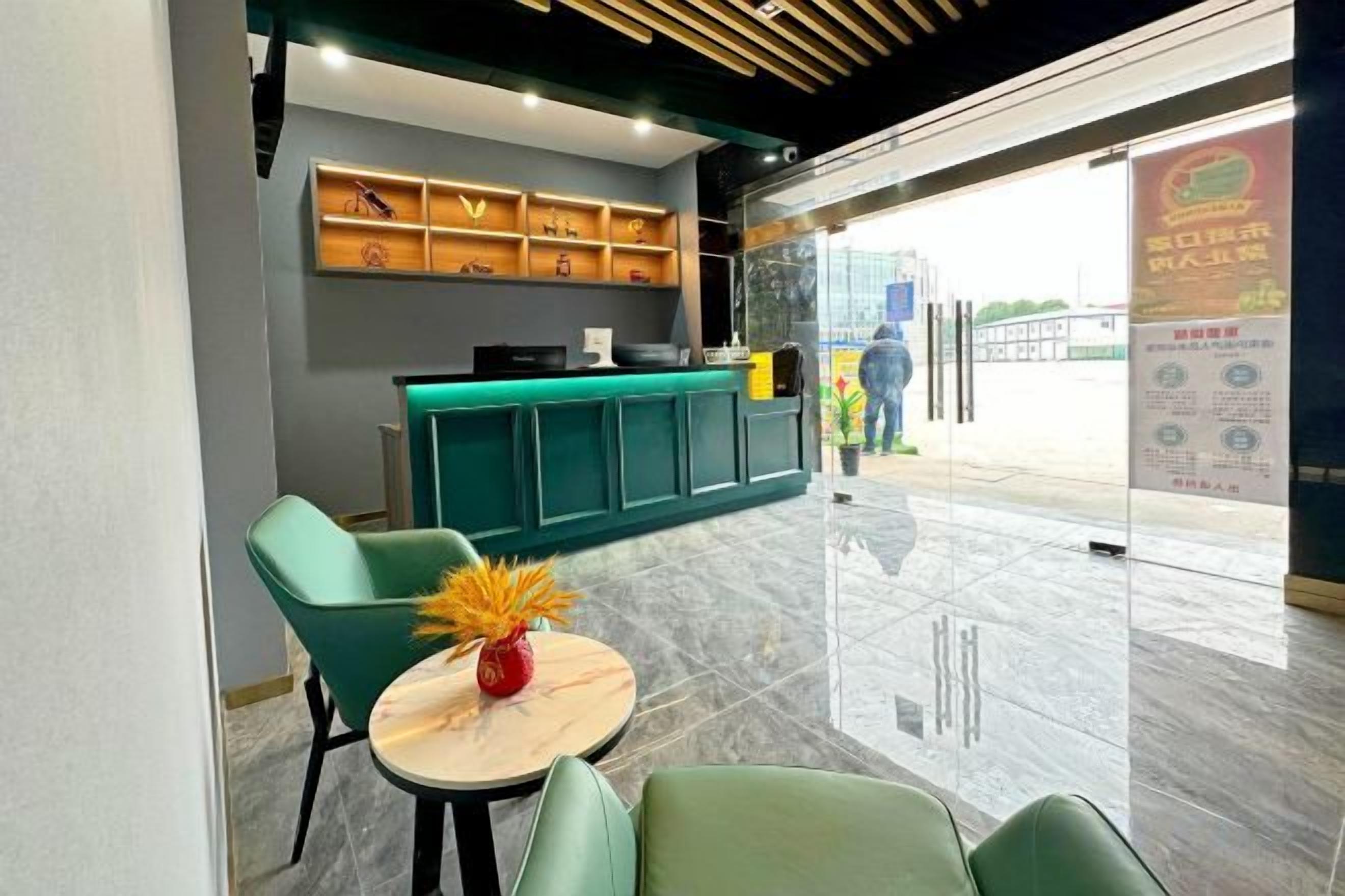 Uma área de recepção com balcão, cadeiras verdes, uma mesa pequena com um vaso e uma porta de vidro que dá para o exterior.