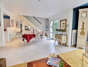 A spacious living area with a dining table set for four, a staircase, and a view of greenery through large windows. - La Maison de Chris - THEIX NOYALO (THEIX NOYALO)