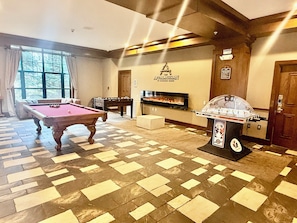 Une table de billard, une table de baby-foot et une cheminée dans une pièce dotée d’un sol à motifs et de poutres en bois.