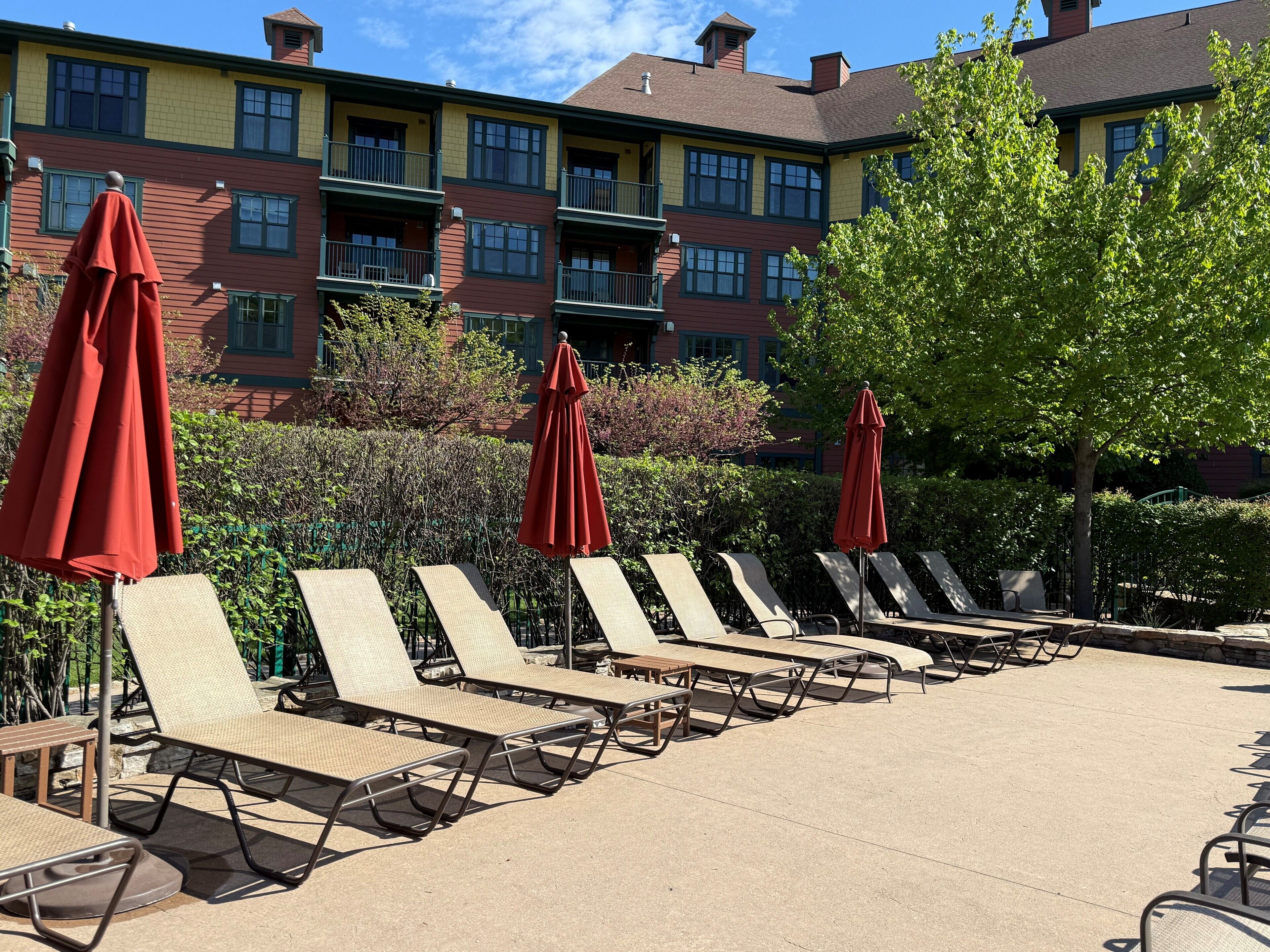 A row of sun loungers with red umbrellas, a stone bench, and a building with multiple balconies in the background.