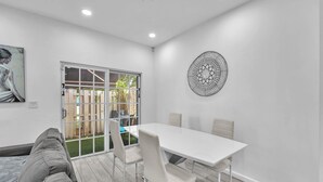 Un espace repas moderne avec une table et des chaises blanches, un canapé gris et un élément décoratif mural.