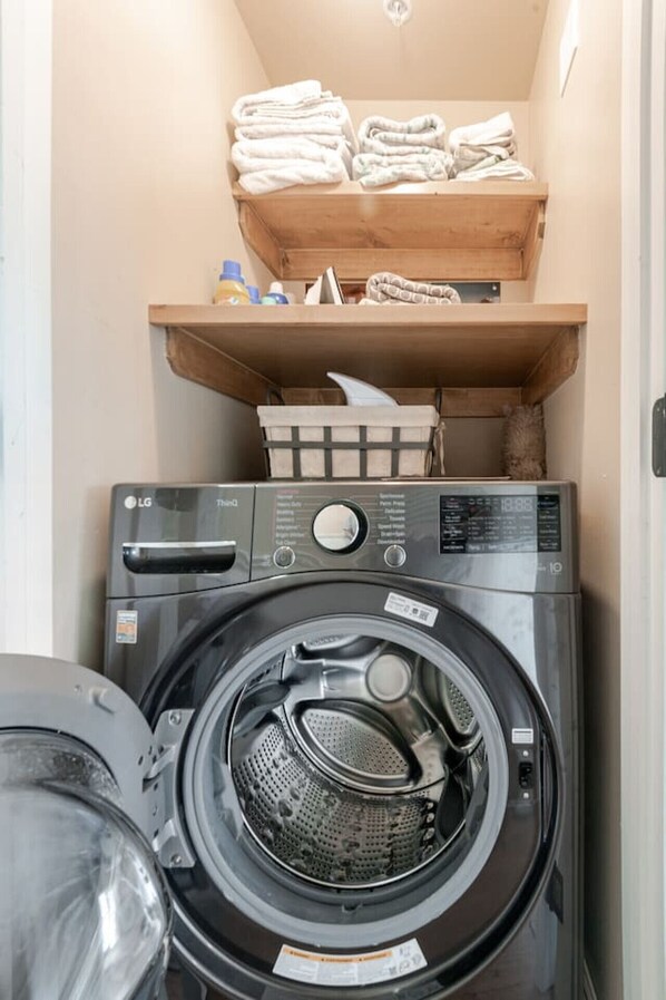 A compact laundry area with a front-loading washing machine, a drying rack, and a shelf with folded towels and toiletries. - Sunset Beach Haven in San Clemente (San Clemente)