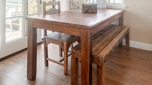 A dining area with a wooden table and bench, large windows with white curtains, and a ceiling light fixture.