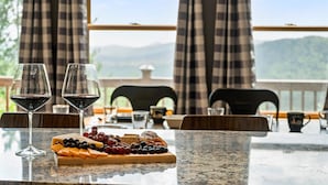 A table with two wine glasses, a cheese board with various cheeses and grapes, and a view of mountains through large windows with checkered curtains.