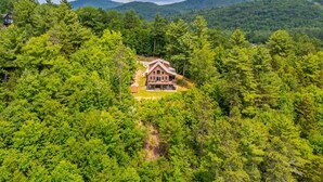 A large house surrounded by dense forest, with a prominent front porch and multiple windows.