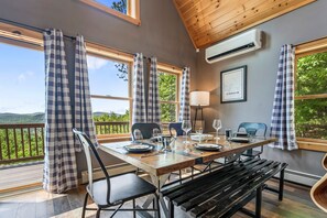 A dining area with a wooden table set for four, large windows with checkered curtains, a wall-mounted air conditioner, and a framed map on the wall. - BryantPond Cabin-BNew Firepit! HotTub|Dog Friendly (Bryant Pond)