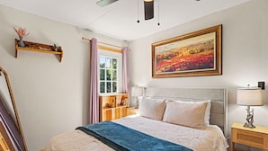 Un dormitorio con cama, mesitas de noche, estante junto a la cama, ventana con cortinas, repisa en la pared y un cuadro.
