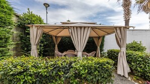 A gazebo with a table and chairs, surrounded by greenery and a white wall. - 1BR Apartment with Tree Views | Golem by PikHost (Golem)