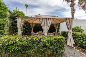A gazebo with a table and chairs, surrounded by greenery and a white wall.