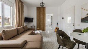 A modern living room with a sofa, a dining table with chairs, a TV mounted on the wall, and a guitar in the background. - 1BR Apartment with Tree Views | Golem by PikHost (Golem)