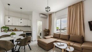 A modern living room with a sofa, a dining table with chairs, a kitchen area with cabinets, and a large window with curtains. - 1BR Apartment with Tree Views | Golem by PikHost (Golem)