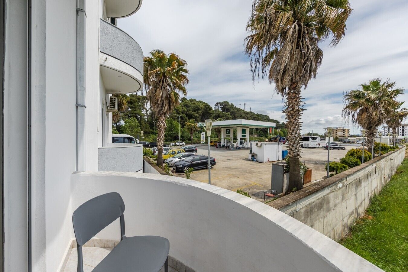 Un balcon avec une chaise, un parking rempli de voitures et, au loin, une station-service.