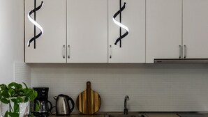 A modern kitchen with white cabinets, a coffee maker, a kettle, and a pizza stone on the counter. There is a white table with chairs and a potted plant. - 1BR Apartment with Tree Views | Golem by PikHost (Golem)