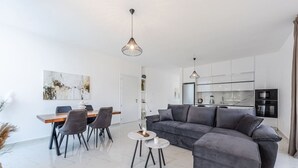 Moderni olohuone, jossa on harmaa kulmasohva, ruokapöytä tuoleineen sekä valkoisilla kaapeilla ja kodinkoneilla varustettu keittiö.
