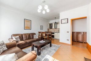 A modern living room with a brown leather sofa, a wooden coffee table, and a dining area with a wooden table and chairs. There is a kitchen visible in the background with white cabinets and a tiled backsplash.