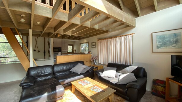 Un vaste espace de vie doté d’un plafond en bois, de deux canapés en cuir noir et d’une table basse en bois, avec une cuisine en arrière-plan.
