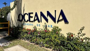 Placa com 'Oceanna Condos de Mar', com boa visibilidade do nome do edifício e área ajardinada com arbustos e flores.