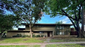 A two-story house with a flat roof, a front porch, and a small front yard with a tree and a pathway leading to the entrance. - Luxury West 7th Fort Worth Stay - Balcony • W/D (Fort Worth)