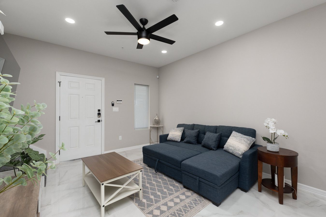 A living room with a dark blue sofa, a wooden coffee table, a ceiling fan, and a small side table with a plant.