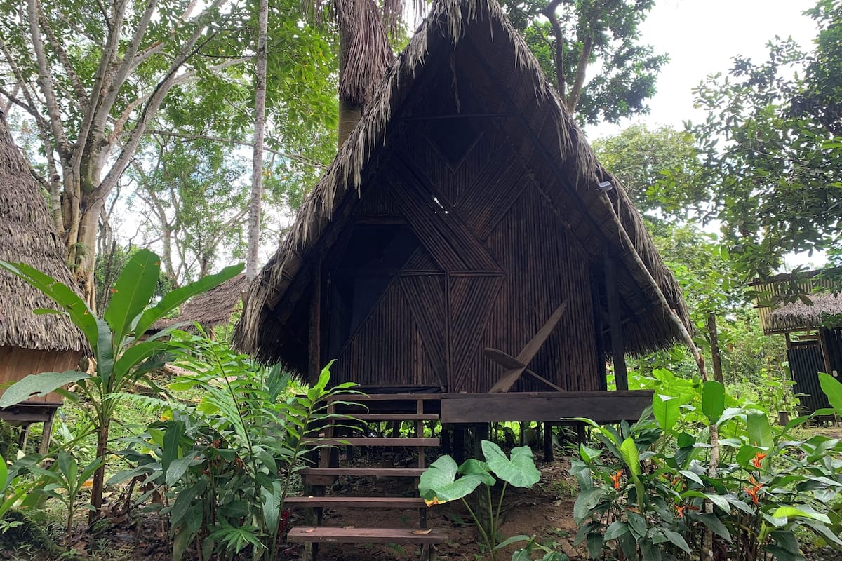 Eine rustikale Hütte mit Strohdach, umgeben von üppiger Vegetation und einer Holztreppe, die zu ihr hinaufführt.