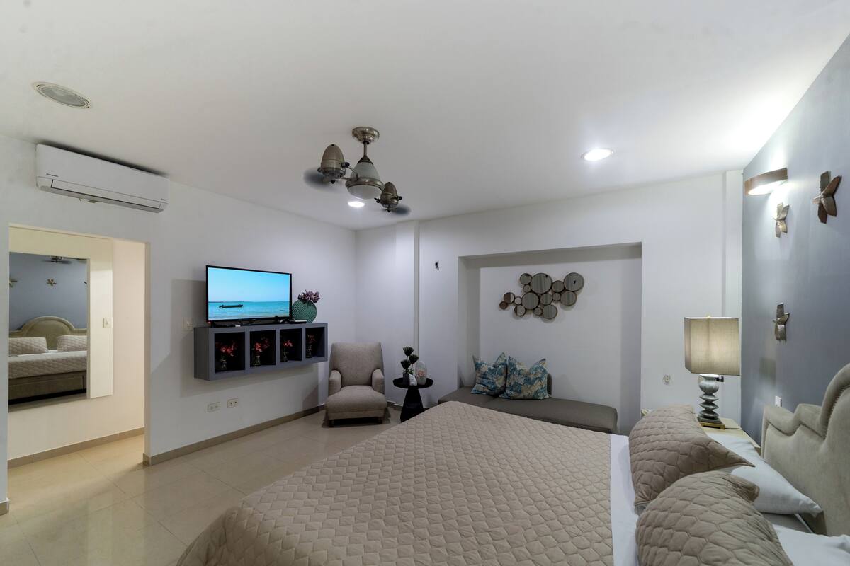 A modern hotel room with a large bed, a TV, a seating area, and a ceiling fan.