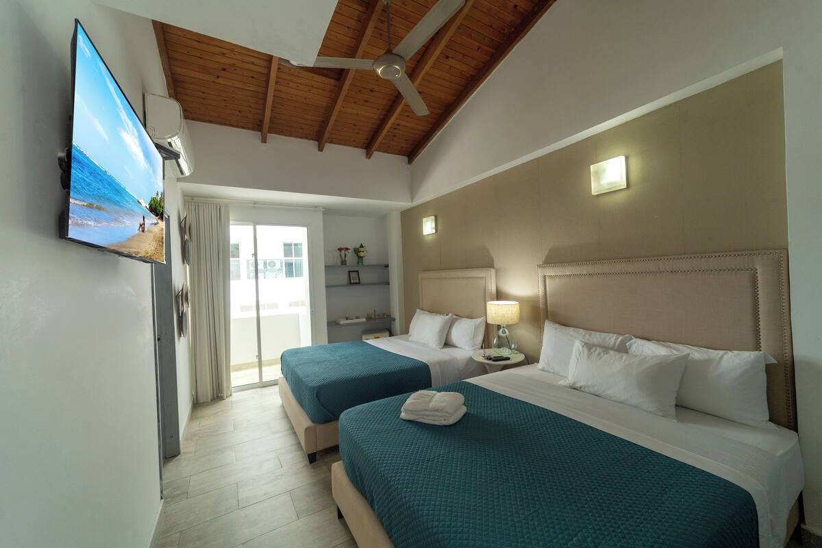 A hotel room with two beds, a ceiling fan, a TV, and a balcony with a view of the beach.